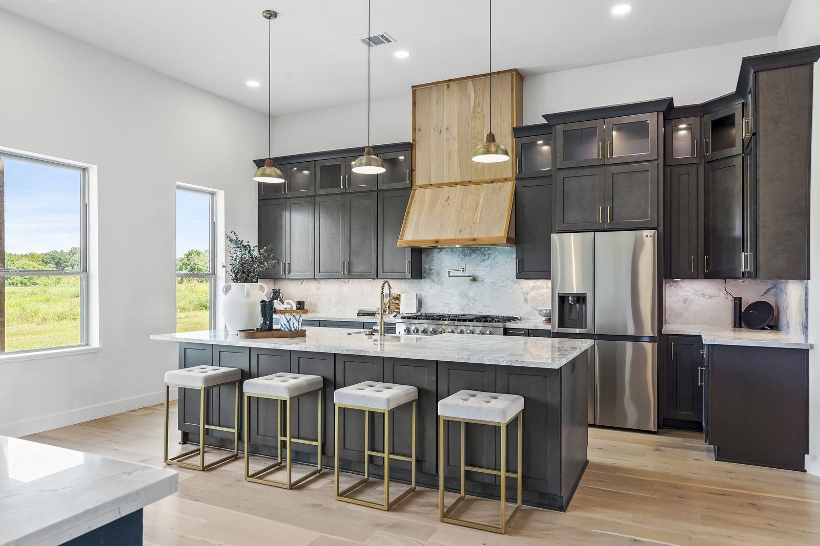 Kitchen with dark shaker cabinets, a large island with counter stools, wood range hood and pendant lights
