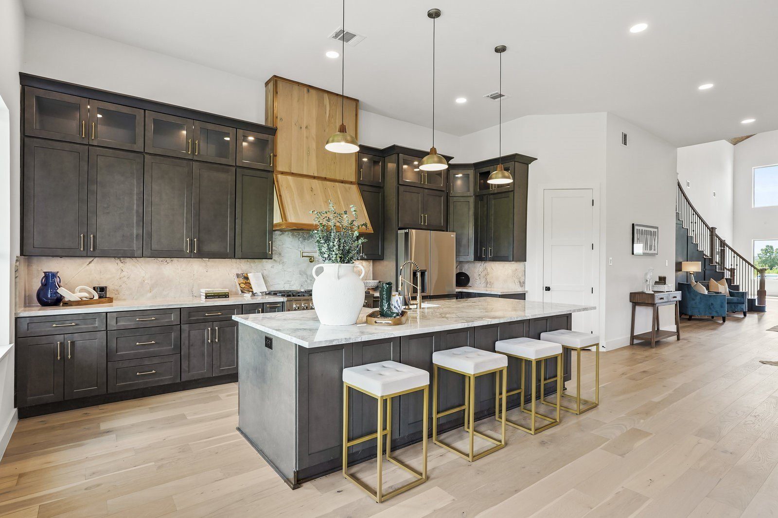 Dark cabinet island with quartz top and gold-base stools, wood range hood and glass-front upper cabinets