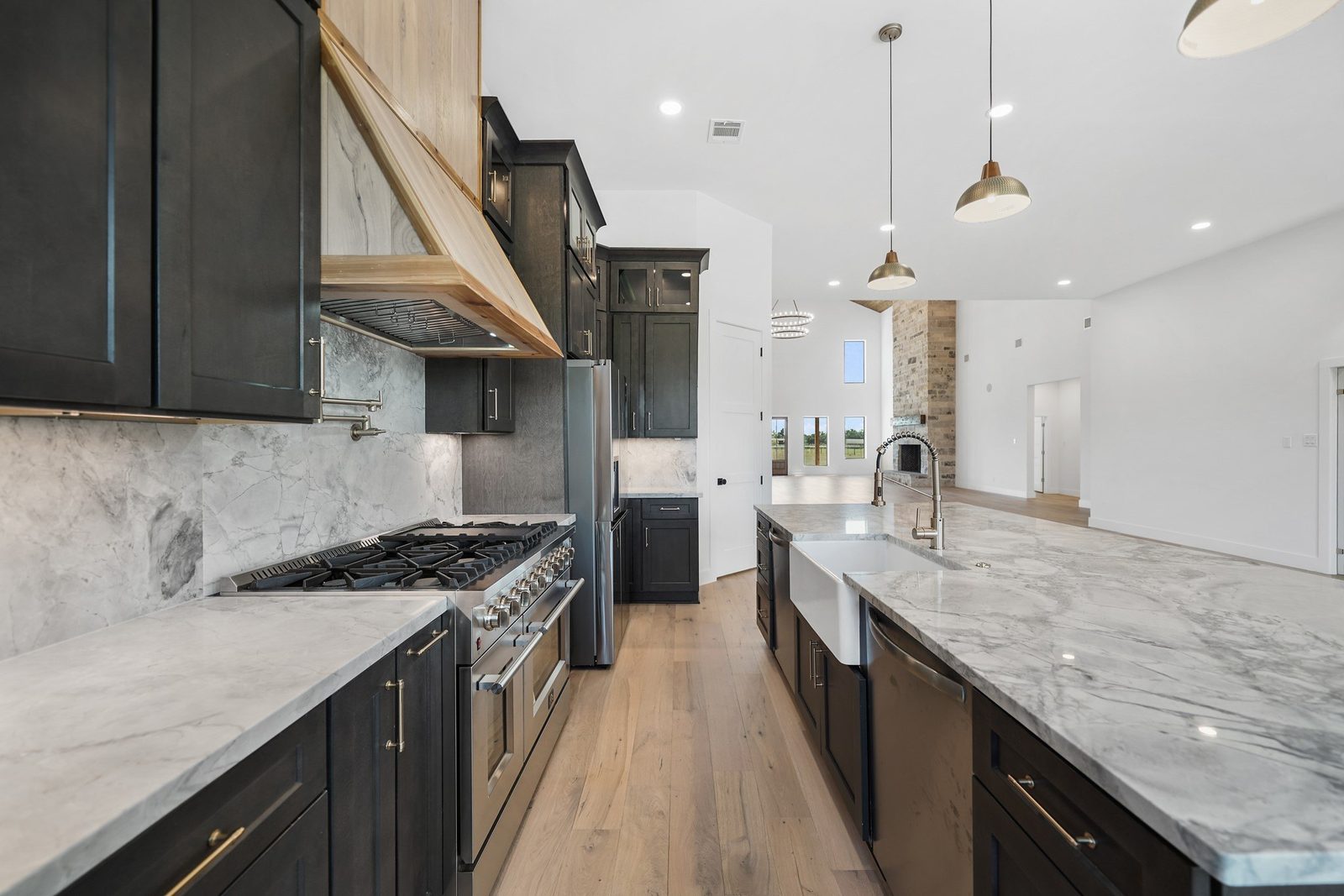 Gas range under a wood hood with a marble-look slab backsplash, farmhouse sink and dark cabinets