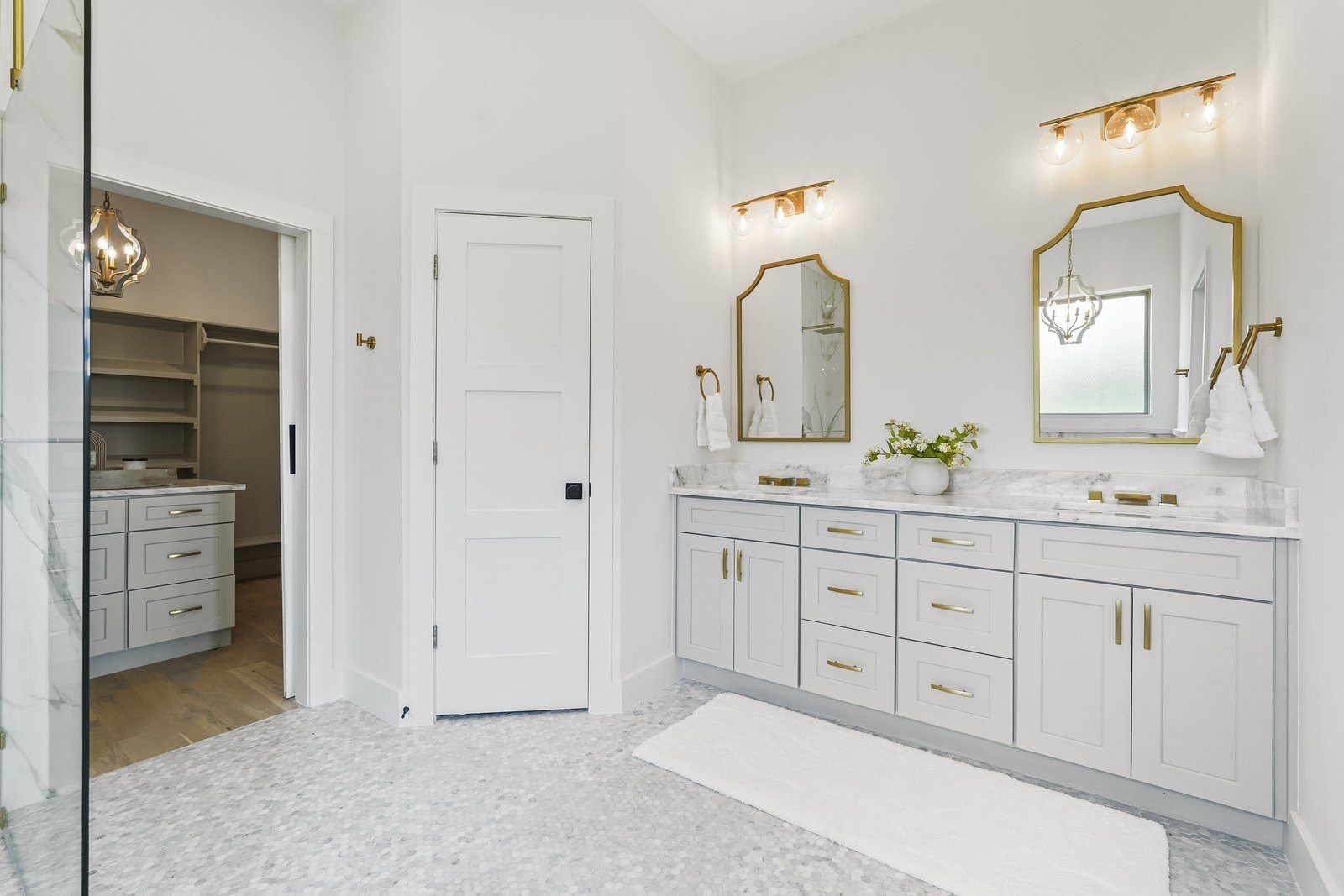 Primary bath with a white double vanity, gold sconces and a doorway to the walk-in closet