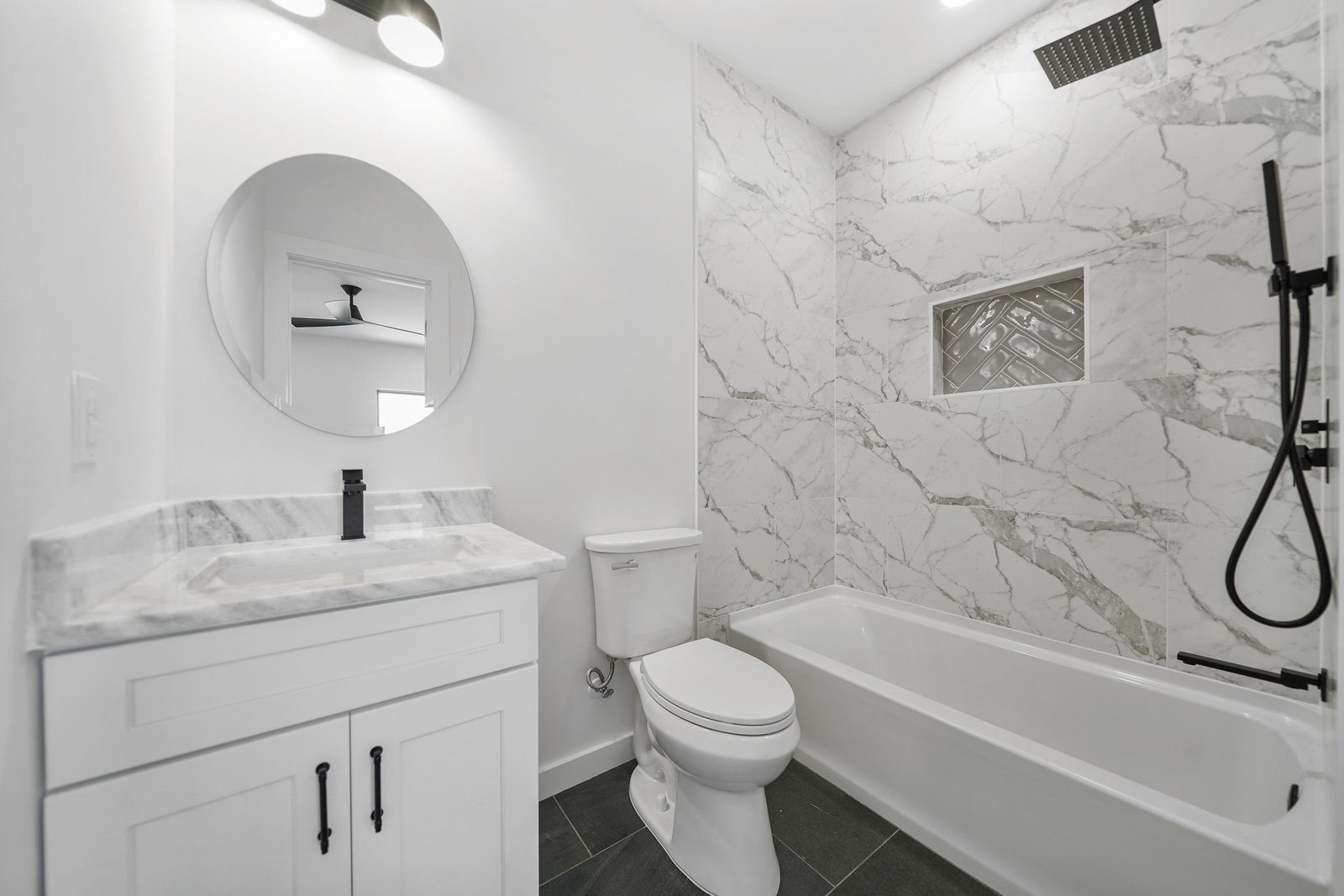 Guest bathroom with a round mirror, white vanity, black fixtures and marble-look tub surround